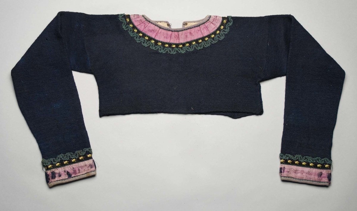 Kort spedetröja i mörkblått ullgarn. Den har använts kring 1860–1880 och är bland annat prydd med anilinlila ylletrikåband och rosa sidenband, som från början varit mycket färgstarka.