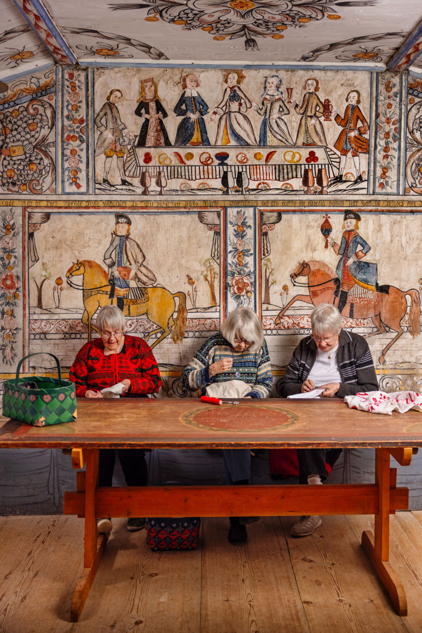 Tre kvinnor sitter framför en väggmålning och broderar.