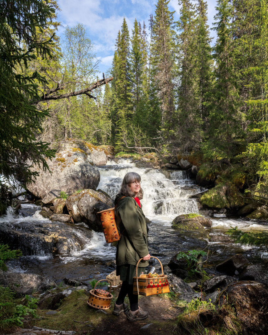 Siv Myhr står vid ett vattenfall med en näverkont på ryggen.
