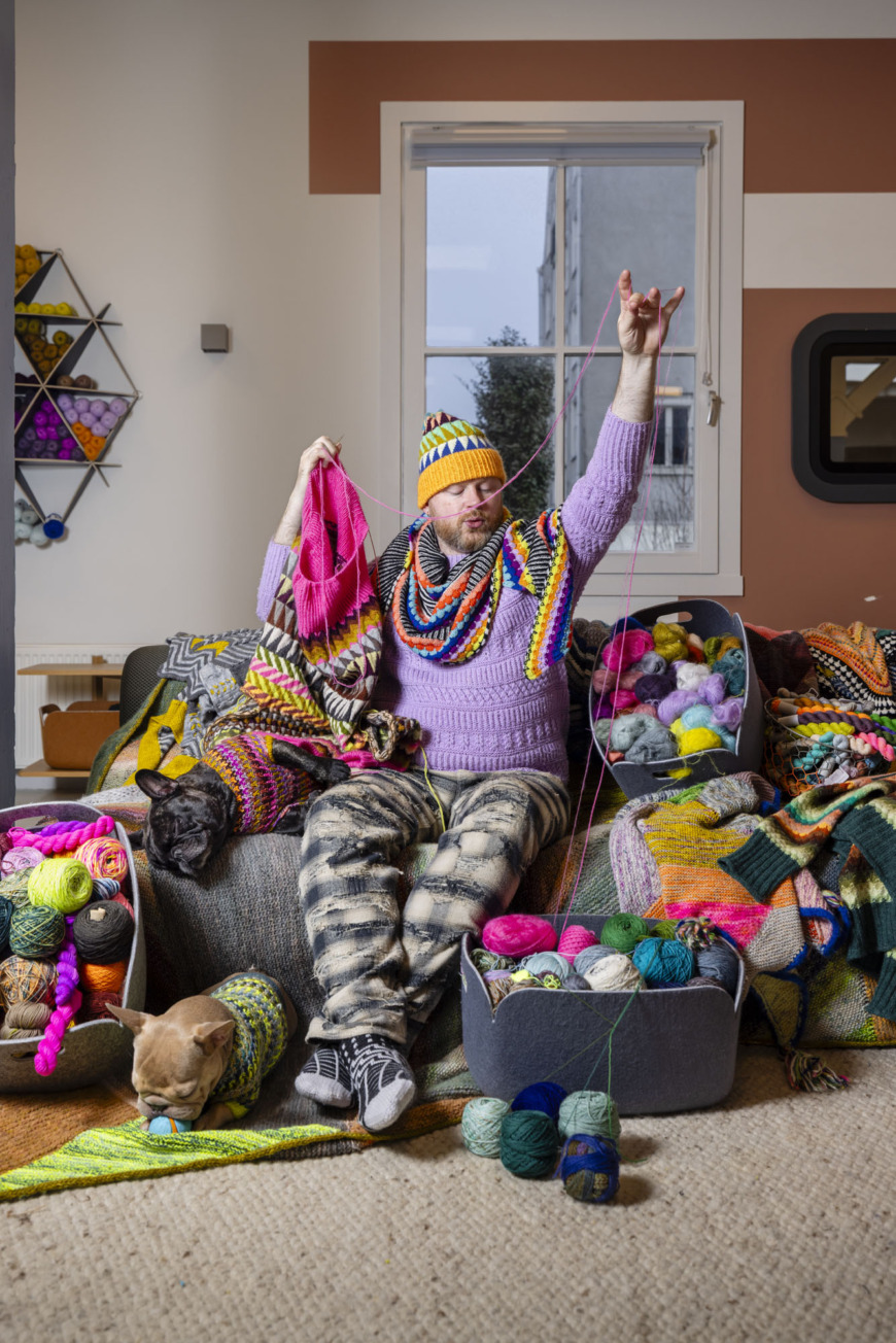 Stephen West sitter i en soffa med många färgglada stickning omkring sig.